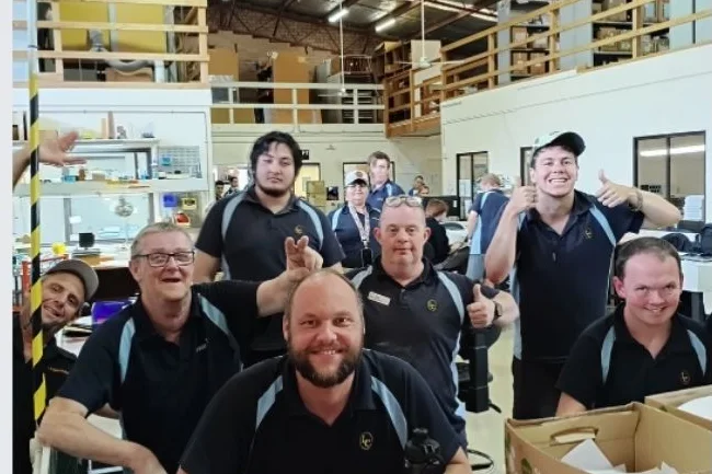 The team at the work benches under the mezzanine at West Gosford, smiling at the camera with cartons of packed orders beside them.
