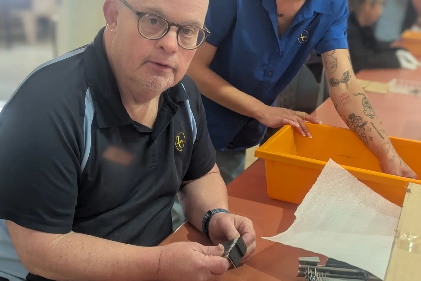 A Lasercraft employee in a branded polo shirt at a bench in the West Gosford workshop, holding a small metal component in both hands, with a colleague reaching into an orange parts tub beside him.