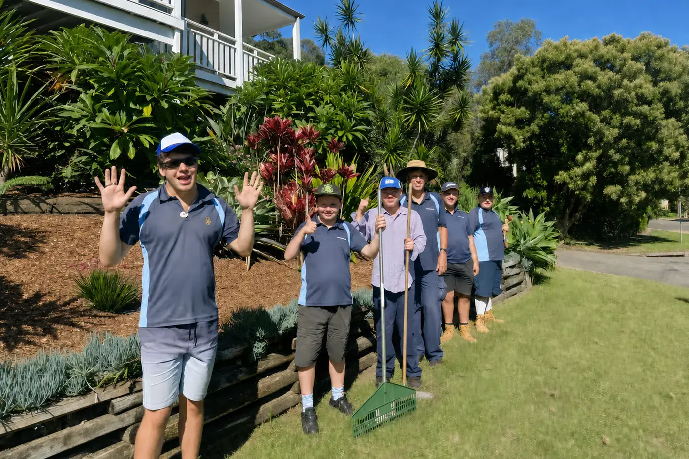 Six members of the Lasercraft gardening crew in matching polo shirts, standing on a mown lawn beside a terraced garden bed in front of a house, two of them waving at the camera and one holding a long handled rake.