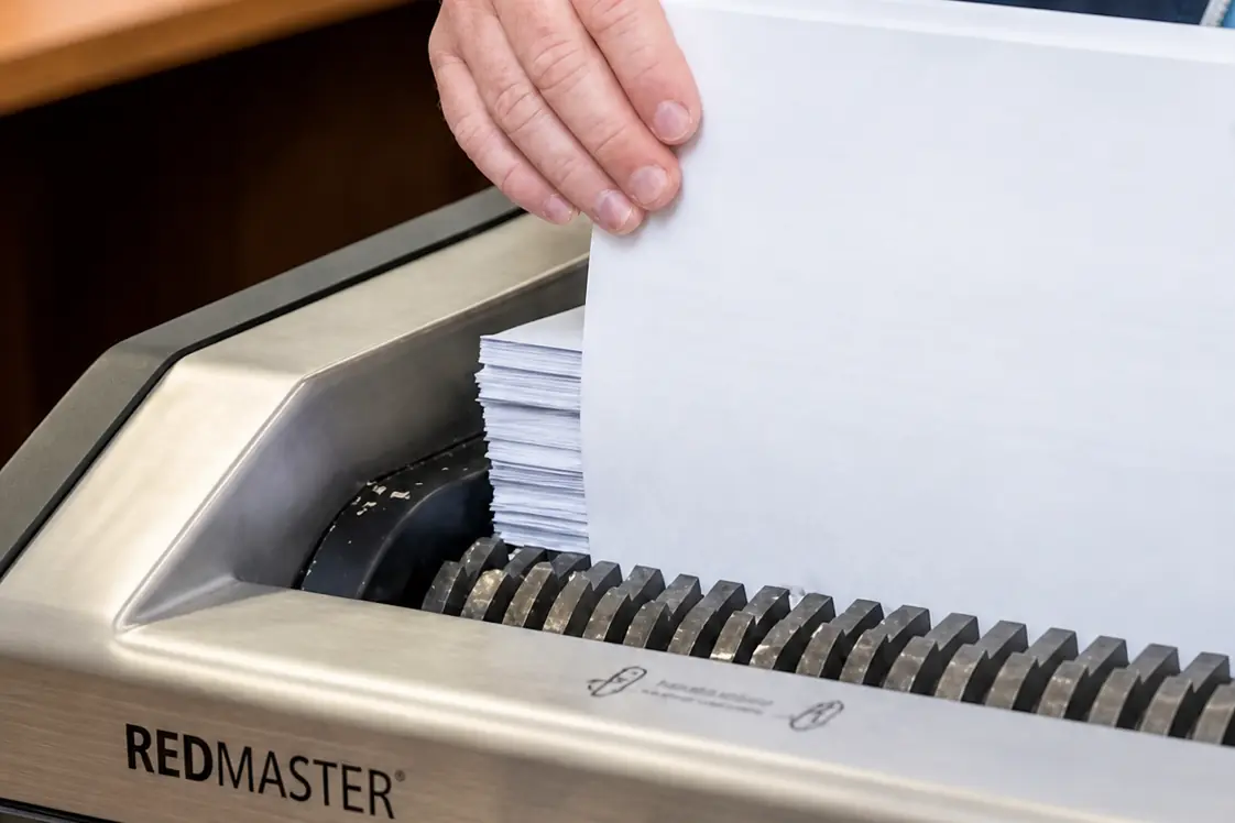 A hand lowering a thick stack of white paper into the toothed cutting head of the industrial shredder at Lasercraft's West Gosford workshop.